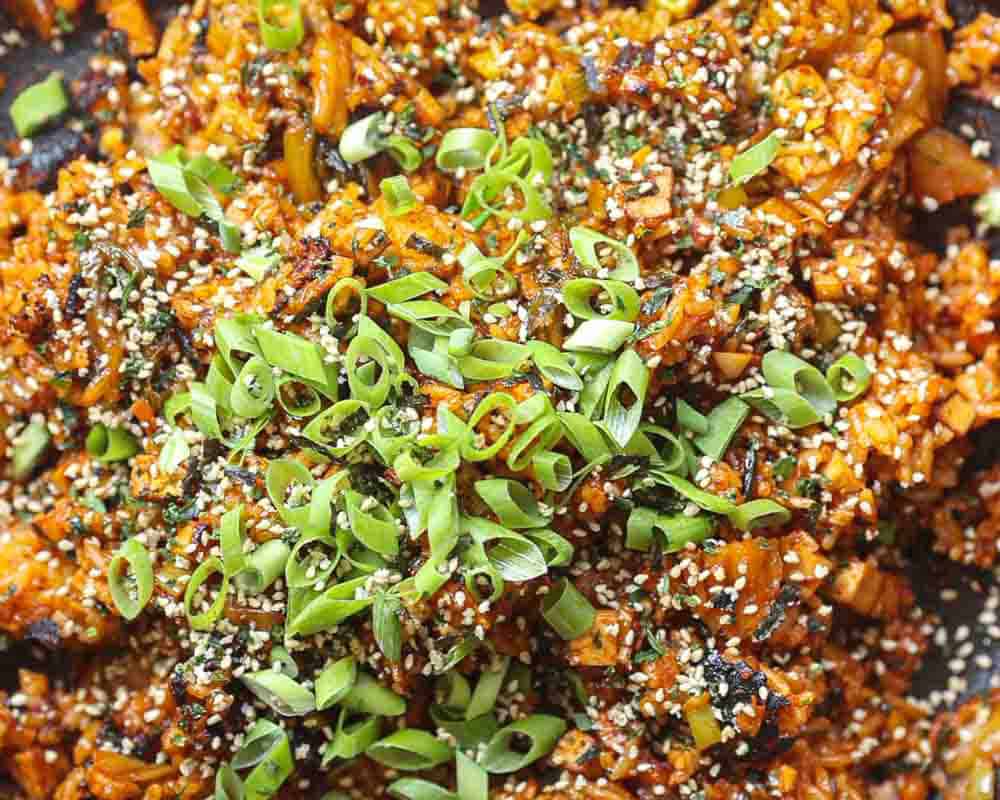 Close-up of a vibrant dish featuring vegan kimchi fried rice mixed with chopped vegetables. The dish is topped with a generous amount of sliced green onions and sprinkled with sesame seeds, giving it a colorful and textured appearance.